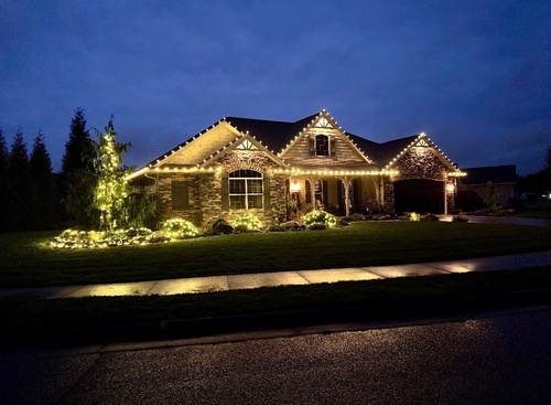 Warm white holiday lights along every roofline of a stone home at dusk, with lit trees and shrubs