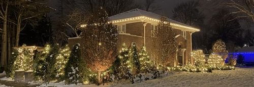 Warm white holiday lighting along a brick home&rsquo;s roofline and wrapped trees in the snow