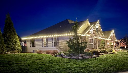 Warm white holiday roofline lighting along a craftsman home&rsquo;s side elevation